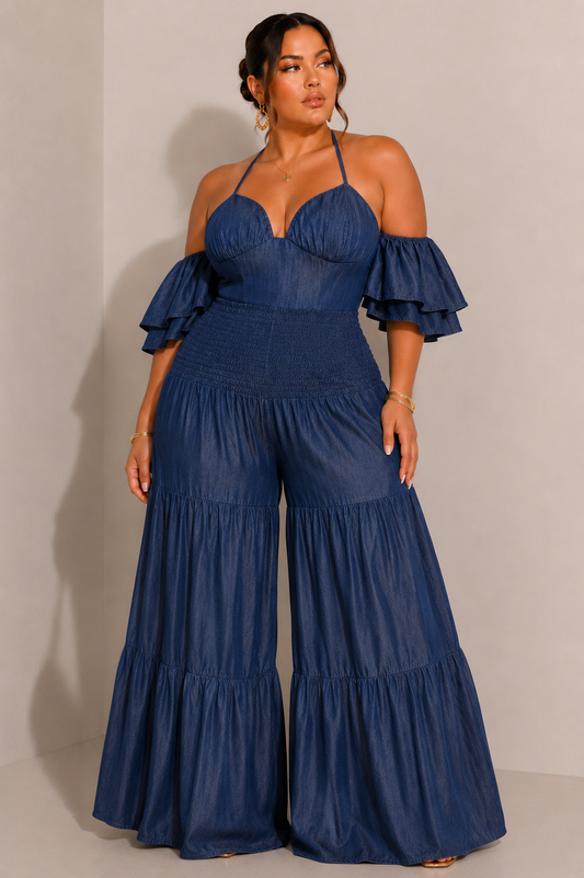Curvy woman wearing a dark chambray halter jumpsuit with off the shoulder ruffle sleeves, a smocked waistband, and wide leg tiered pants, styled for brunch, vacation, or date night.