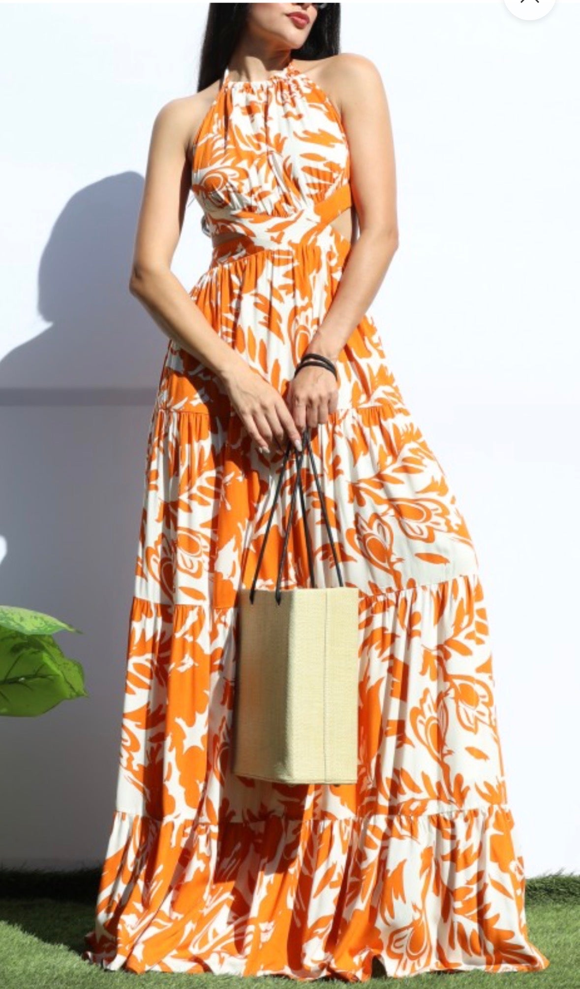 Woman wearing an orange and ivory tropical print halter maxi dress with an open back and tiered skirt, styled with a straw tote bag for a summer brunch or vacation look.