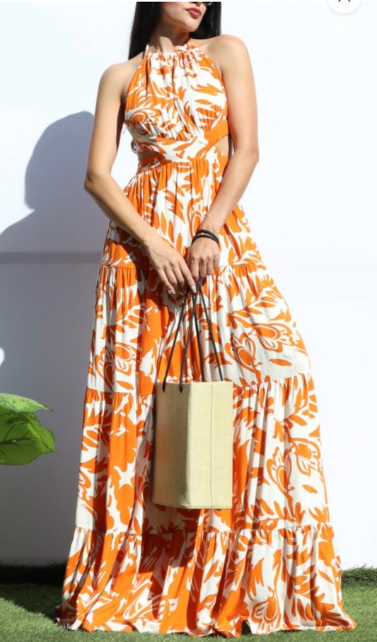 Woman wearing an orange and ivory tropical print halter maxi dress with an open back and tiered skirt, styled with a straw tote bag for a summer brunch or vacation look.